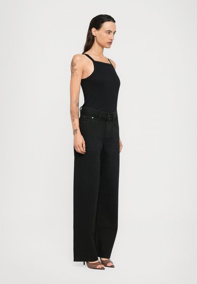 Woman wearing a black sleeveless top, high-waisted wide-leg black jeans, and brown open-toe heels, standing sideways on a white background.