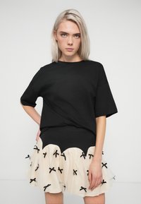 Jovem mulher com cabelo platinado e louro, vestindo uma t-shirt preta larga e uma saia curta creme decorada com pequenos laços pretos.