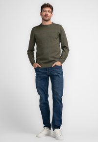 Olijfgroene gebreide trui met ronde hals, gecombineerd met blauwe spijkerbroek en witte sneakers. De pasvorm is casual en ontspannen.