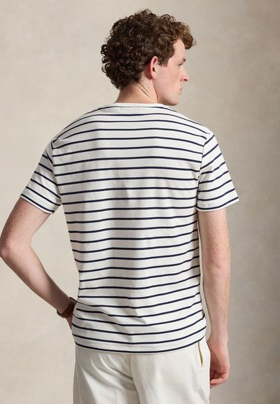 Νεαρός άνδρας με σγουρά μαλλιά, φορώντας λευκό κοντομάνικο πουκάμισο με οριζόντιες ναυτικές ρίγες, στέκεται με το ένα χέρι στη μέση του.