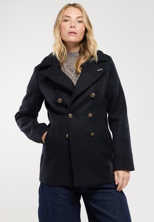 Cappotto peacoat doppiopetto blu navy con grandi bottoni di bronzo, ampie rever e un maglione a maglia testurizzata sotto. Presenta tasche laterali.