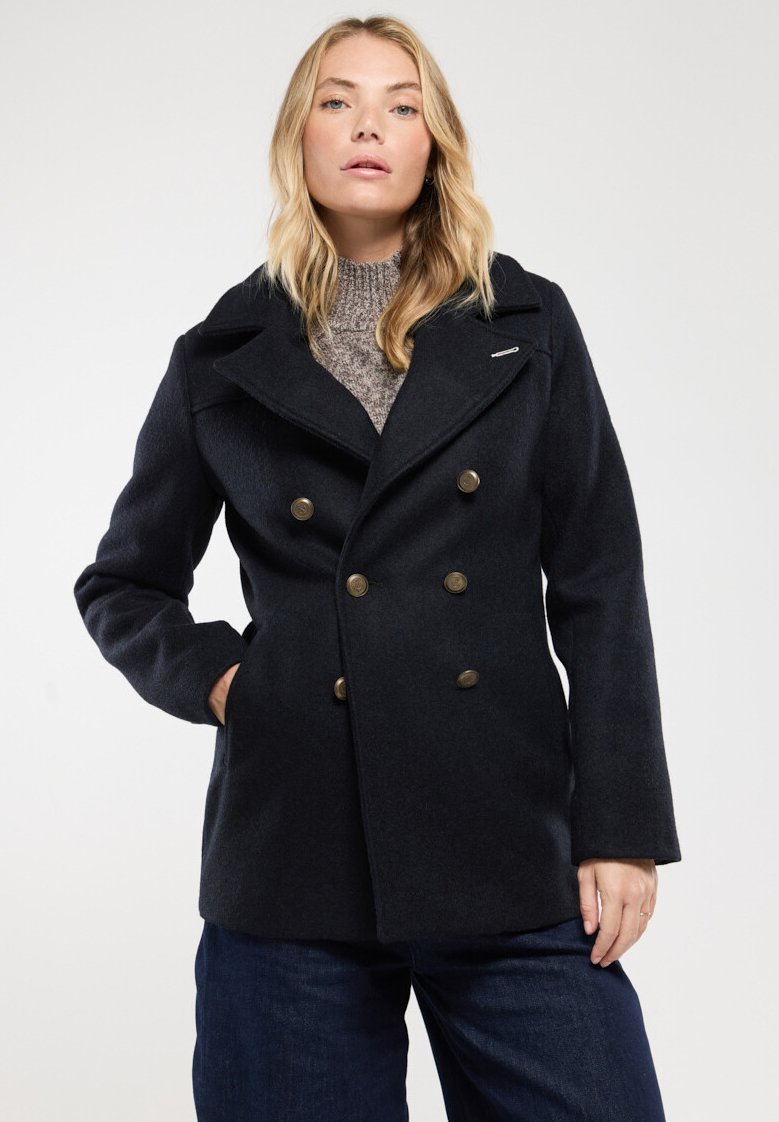 Abrigo de marina de doble botonadura con grandes botones de bronce, solapas anchas y un suéter de punto texturizado debajo. Cuenta con bolsillos laterales.