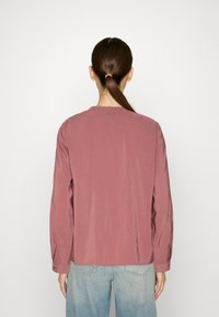 Maglietta a maniche lunghe in tessuto rosa polveroso, caratterizzata da un collo rotondo con bordatura arricciata, abbinata a jeans blu chiaro sbiaditi. Design semplice e casual.