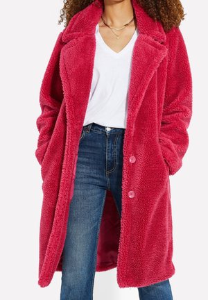 Kobieta w długim, jaskraworóżowym, puszystym płaszczu na białej koszulce z dekoltem w serek i niebieskich dżinsach, z rękami w kieszeniach płaszcza.