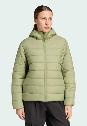 Junge Frau trägt eine hellgrüne Steppjacke mit Kapuze, steht vor einem schlichten hellen Hintergrund und schaut zur Seite.