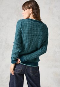 Pull en tricot teal avec un motif texturé, ourlet et poignets côtelés, porté sur un jean en denim foncé ; la vue de dos montre les détails des épaules et de l'encolure.