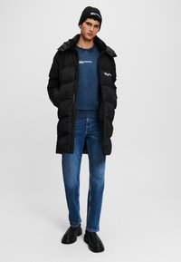 Palton puf negru cu glugă, pulover tricotat albastru, blugi din denim și cizme negre. Puloverul are un logo alb, iar țesătura sa este vizibilă.