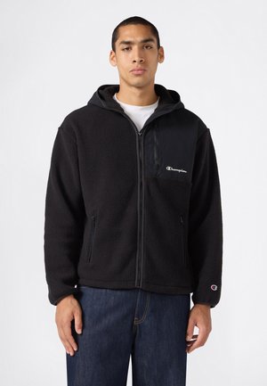 Junger Mann trägt eine schwarze Fleecejacke mit Reißverschluss und Kapuze, Champion-Logos auf Brust und Ärmel, und dunkelblaue Jeans vor weißem Hintergrund.