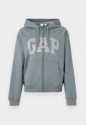 Sweat à capuche gris zippé avec poches avant et capuche, arborant le mot "GAP" en grandes strass blancs sur la poitrine.