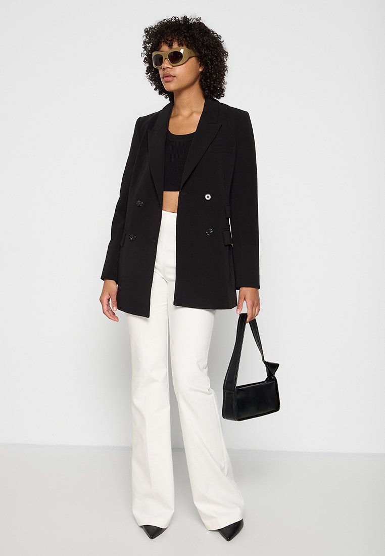 Schwarzer, doppelt geknöpfter Blazer mit Revers, kombiniert mit einem schwarzen Crop-Top und weißen, ausladenden Hosen. Accessoires: kleine schwarze Tasche und Sonnenbrille.