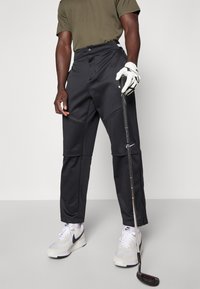 Black golf pants made of stretchy fabric, featuring side panels, a tailored fit, and a zippered hem, paired with white sneakers and a golf club.