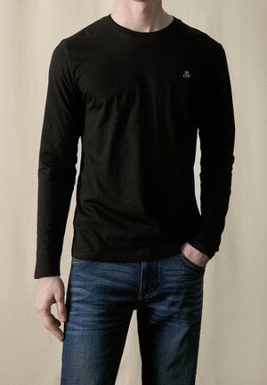 Mężczyzna w dopasowanej czarnej koszulce z długim rękawem z małym białym logo "M OP" na piersi i w niebieskich dżinsach, jedną ręką w kieszeni.