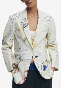 Blazer blanc avec des contours floraux colorés en bleu, jaune et marron. Il possède un design ajusté, des revers en pointe et deux boutons sur le devant.