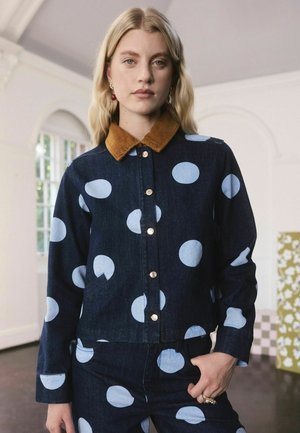 Blondīne sieviete, kas valkā tumši zilu džinsa žaketi un bikses ar lieliem gaiši ziliem punktiņiem, stāv iekštelpās ar roku kabatā.