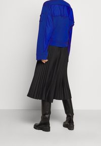 Persona vistiendo una chaqueta azul, una falda midi plisada negra y botas negras hasta la rodilla, de pie contra un fondo claro liso.