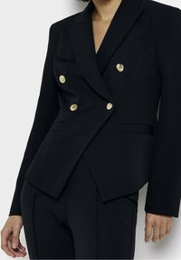 Kvinna iklädd en skräddarsydd svart blazer med sex gula knappar och matchande svarta byxor, stående mot en vit bakgrund.