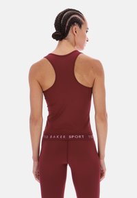 Bordó sportos ujjatlan felső versenyhátsó kialakítással és sima textúrával. Márkázott derékszíj jellemzi, amelyen "TED BAKER SPORT" felirat található kontrasztos színben.