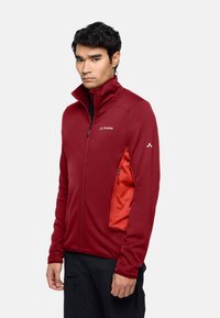 Rote Zip-Jacke mit hohem Kragen, ausgestattet mit einer kontrastierenden orangefarbenen Seitentasche, strukturiertem Stoff und schwarzem Markenlogo auf der Brust.