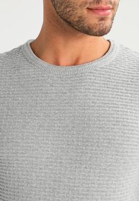 Homme avec une légère barbe portant un pull à col ras du cou gris clair en laine texturée, visible du cou à la poitrine sur un fond uni.