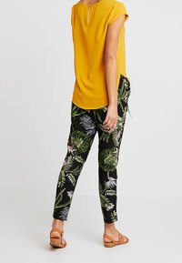Persona che indossa una maglietta gialla a maniche corte e pantaloni neri con motivi tropicali di foglie verdi e rosa, abbinati a sandali marroni, in piedi.