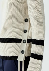 Strickpullover in Creme mit schwarzen horizontalen Streifen. Verfügt über eine seitliche Öffnung mit fünf großen schwarzen Knöpfen und geripptem Textil.