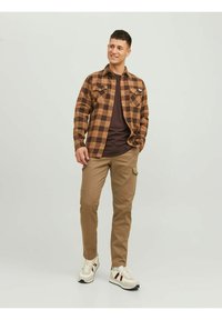 Camicia a quadri marrone con design button-up, abbinata a pantaloni cargo color khaki e sneakers bianche con accenti rossi. Stile casual per un look quotidiano.