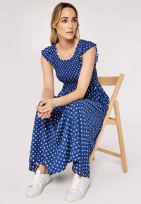 Blue polka dot maxi dress with short ruffled sleeves, smocked bodice, and a flowing skirt. Paired with white sneakers. Wooden chair in background.
