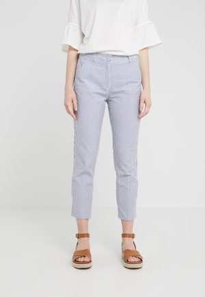 Personne portant un chemisier blanc à manches volantées, un pantalon court à rayures verticales bleu et blanc, et des sandales marron à brides autour de la cheville.