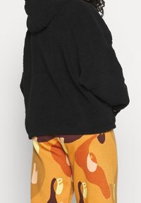 Sudadera de forro polar negra con una superficie texturizada, combinada con leggings de patrones naranjas y amarillos que presentan formas y curvas abstractas.