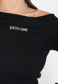 Musta ribbed off-shoulder neulepusero, jossa on valkoisella brodeerattu "SIXTH JUNE" etuosassa. Pehmeä rakenne ja istuva muotoilu.