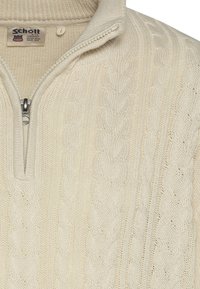 Jasnokremowy sweter dzianinowy z zamkiem na ćwiartkę przy kołnierzu, ozdobiony wzorami warkoczowymi i prążkowanymi detalami na szyi oraz dole.