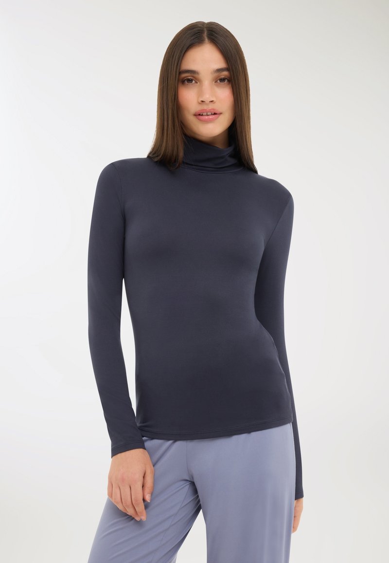 Kobieta z prostymi brązowymi włosami, ubrana w dopasowany ciemnoszary golf i luźne jasnoszare spodnie, stojąca na jednolitym jasnym tle.