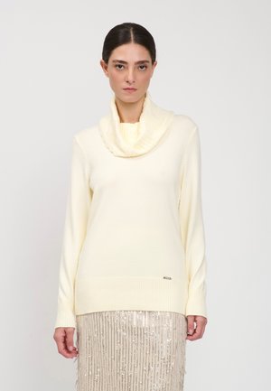 Donna con capelli scuri che indossa un maglione crema con collo ad anello e una gonna beige con paillettes, in piedi davanti a uno sfondo bianco semplice.