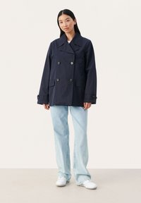 Mørkeblå dobbeltbrystet jakke med store knapper, sidelommer og bred krave, parret med lyseblå brede jeans og hvide sneakers.