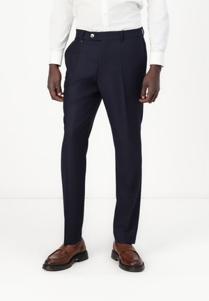 Sötétkék nadrág, sima anyagból, slim fit szabással, elöl redőzött és gombos záródással, barna bőr loafer cipőkkel párosítva.
