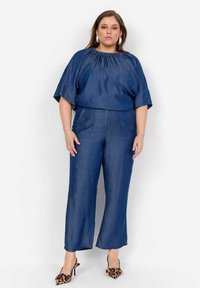 Denimoutfit som inkluderar en topp med samlad hals och vida byxor i mörkblått, med fickor och en slät textur, kombinerat med klackar i leopardmönster.