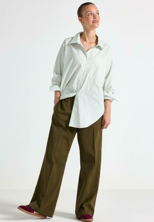 Camicia button-up a righe verde chiaro e bianco con maniche arrotolate, abbinata a pantaloni larghi verde oliva e scarpe slip-on bordeaux.