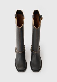 Botas negras altas con acentos marrones, exterior suave, suelas planas y correas laterales con hebillas. Forro interior texturizado visible en la parte superior.