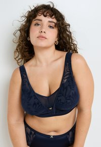 Soutien-gorge bleu marine avec un design en dentelle florale, support avec armatures, bretelles réglables et une bande lisse. Présente un décolleté en V et une texture en double tissu.