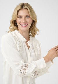 Blouse blanche avec des détails en dentelle sur les épaules et les poignets. Elle a un col à volants et une fermeture à boutons, fabriquée en tissu léger.
