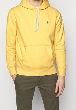 Mann trägt einen gelben Hoodie mit Fronttasche und blauem Logo, kombiniert mit dunkelgrüner Hose, steht mit entspannten Händen.