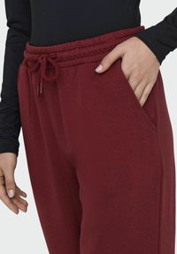 Pantalones de chándal burdeos con cintura elástica y cordón; cuenta con bolsillos laterales y un sutil detalle bordado en el muslo. Textura suave.
