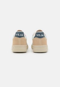 Ett par beiga och pastellfärgade Veja-sneakers visade från baksidan med vit logotyp på marinblå hälflik mot vit bakgrund.