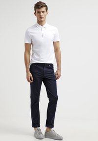 Weißes Kurzarm-Polohemd mit einem Drei-Knopf-Verschluss, kombiniert mit schmal geschnittenen dunkelblauen Hosen und hellgrauen Sneakers.