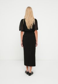 Schwarzes Kleid mit kurzen Puffärmeln, elastischer Taille und gerader Silhouette. Glatter Stoff mit zwei Gesäßtaschen und einem Schlitz. Schwarze Ballerinas getragen.