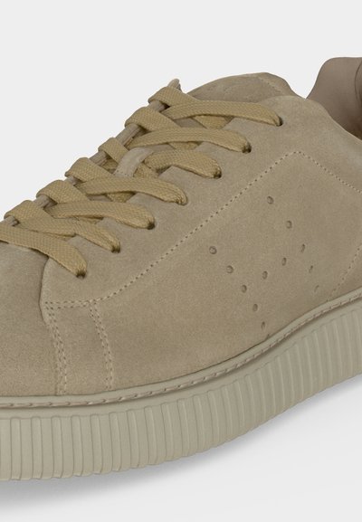 Baskets en daim beige avec des lacets assortis et une semelle en caoutchouc rainurée, présentant de petits motifs perforés de trous sur le côté.