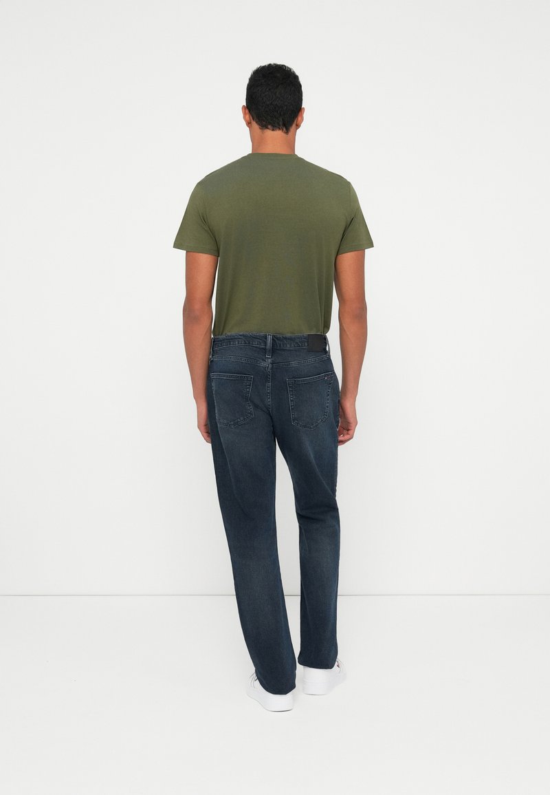 Olive green short-sleeve T-shirt paired with dark blue jeans and white sneakers, viewed from the back, showcasing fit and style details.