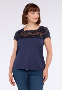 Donna con capelli castani corti che indossa un top blu navy con scollatura in pizzo e maniche corte, abbinato a jeans blu, su uno sfondo bianco.