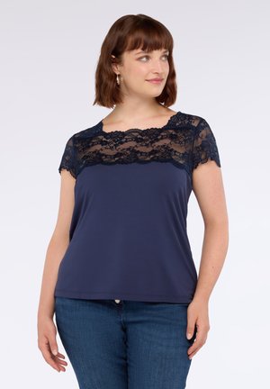 Donna con capelli castani corti che indossa un top blu navy con scollatura in pizzo e maniche corte, abbinato a jeans blu, su uno sfondo bianco.