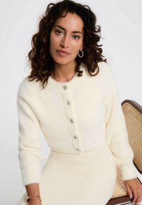 Cardigan morbido e soffice di colore crema con cinque bottoni blasonati, scollatura tonda e polsini a costine, abbinato a una gonna a costine coordinata.